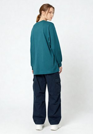 Femme debout regardant en arrière, portant un haut à manches longues turquoise, un pantalon cargo bleu marine et des baskets blanches sur un fond blanc uni.