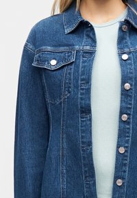 Γυναίκα που φοράει ένα μεσαίου μπλε χρώματος τζιν μπουφάν με ασημένια κουμπιά πάνω από ένα ανοιχτό μπλε ριμπ τοπ, το οποίο είναι κομμένο στους ώμους και στο σώμα.