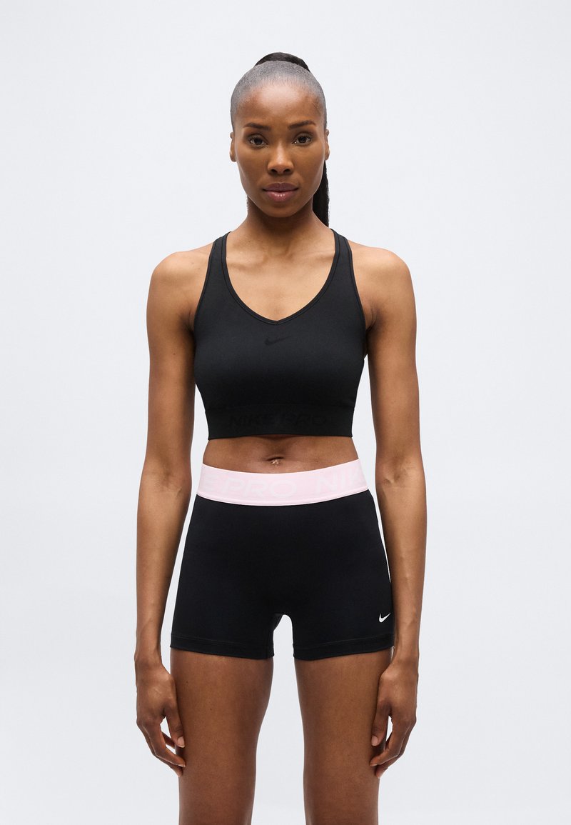 Femme en forme debout, portant un soutien-gorge de sport noir et un short noir avec une ceinture rose, sur fond blanc uni.