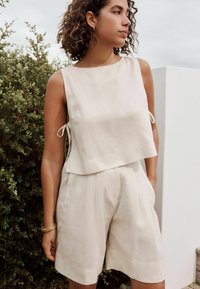 Ensemble deux pièces en lin beige clair comprenant un haut sans manches avec des liens sur les côtés et un short ample avec poches, sur fond de verdure.