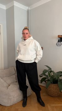 Sweatshirt branca com capuz e logótipo, combinada com calças pretas largas e botins pretos. Ao fundo, uma planta verde e um sofá texturizado.