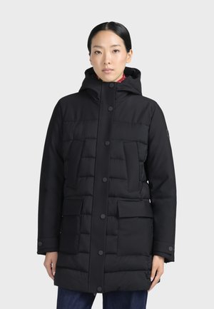 Femme portant un manteau matelassé noir à capuche avec des boutons-pression à l'avant et plusieurs poches, debout devant un fond clair uni.