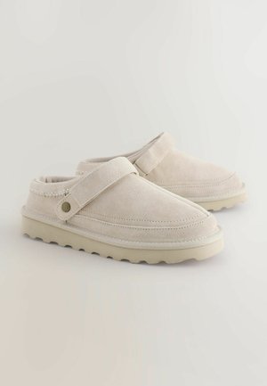 Chaussures plates en daim beige à enfiler, avec une semelle en caoutchouc plate, une large bride et des surpiqûres le long de la tige.