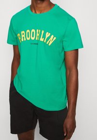 Camiseta de algodón verde con cuello redondo, con la palabra "BROOKLYN" en texto amarillo arqueado y un logo negro debajo. Llevada con pantalones cortos negros.