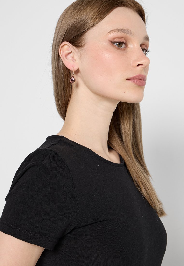 Goldfarbene Ohrringe mit Tropfen-design, die einen birnenförmigen rosa Edelstein zeigen, getragen von einem Modell mit glattem braunem Haar. Schwarzes Shirt.