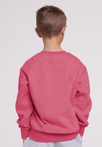 Bambino con i capelli corti castani che indossa una felpa rossa e pantaloni grigi, girato di spalle contro uno sfondo chiaro e semplice.