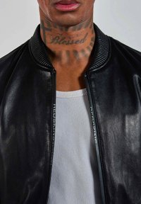 Veste bomber en cuir noir avec col côtelé, dotée d'une fermeture éclair et d'un logo le long de la fermeture; portée par-dessus un t-shirt côtelé gris clair.