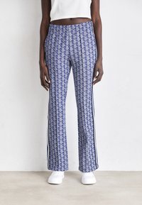 Person wearing blue patterned wide-leg pants with white side stripes and white sneakers, standing against a plain light wall.