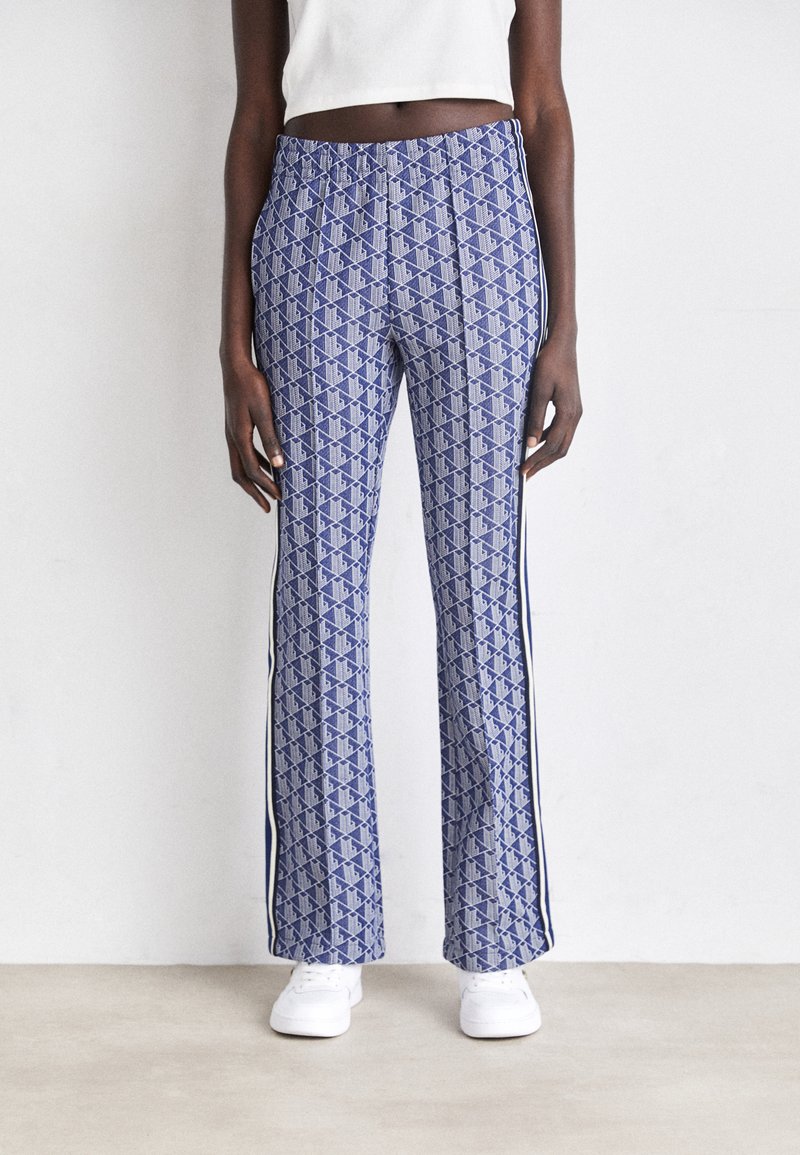 Person wearing blue patterned wide-leg pants with white side stripes and white sneakers, standing against a plain light wall.