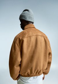 Veste en daim marron avec un col haut, coupe ample, ceinture élastique et bonnet en tricot gris porté sur la tête. Texture lisse, détails minimaux.