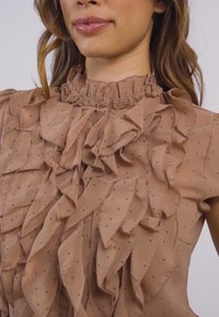 Beige ruffled blůza s vysokým límcem, zdobená puntíkovými akcenty. Tkanina má měkkou, hladkou texturu a vrstvené volány po celé ploše.