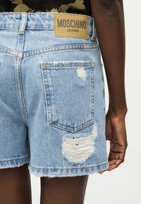 Ανοιχτό μπλε σορτς από τζιν με φθορές, με πίσω τσέπη και ταμπέλα Moschino Jeans σε ταμπά χρώμα, φορεμένα από άτομο με σκούρο δέρμα.