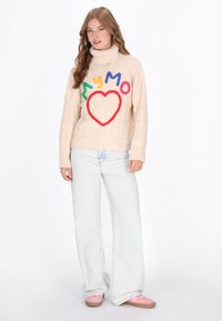 Beige gestrickter Pullover mit einem Rollkragen, der bunte Buchstaben und ein Herzdesign zeigt. Kombiniert mit hellblauen weiten Jeans und rosa Schuhen.