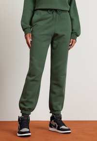 Gröna bomullssvettsbyxor med elastiska fållar och dragsko i midjan, tillsammans med svarta och grå höga sneakers. Slät textur, avslappnad design.