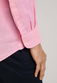 Camisa de lino rosa claro con puños abotonados y un corte relajado, combinada con pantalones de color oscuro. Enfocándose en la textura y la tela suave.