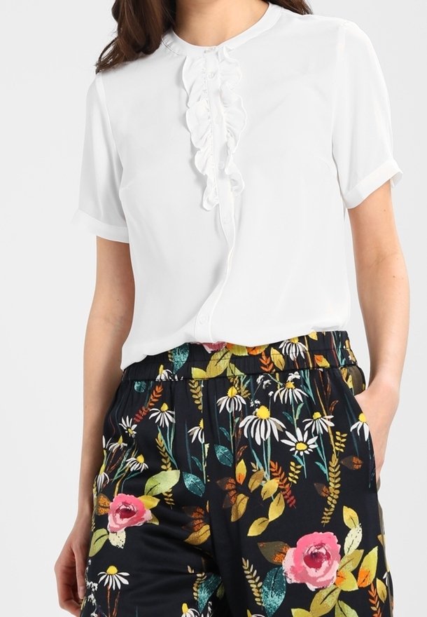Femme portant une blouse blanche à manches courtes avec des volants à l'avant, associée à un pantalon noir avec des imprimés floraux colorés.