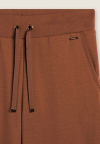 Pantalons de survêtement marron avec une taille à cordon de serrage et des embouts métalliques, texture douce et branding minimal près de la taille.