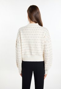 Teksturowany sweter w kolorze kremowym z małymi, beżowymi kropkami, z prążkowanymi mankietami i dołem. Krótki fason z luźnym krojem. Noszony na czarnych spodniach.