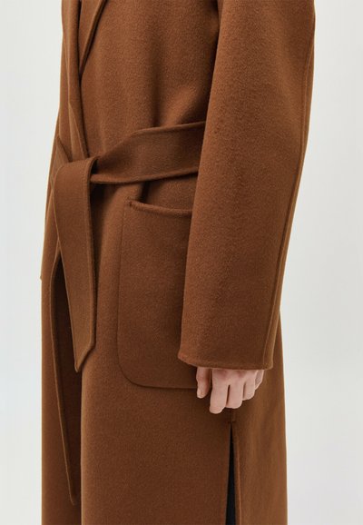 Palton maro din lână cu o curea legată în talie și un buzunar mare în față, prezentat pe partea dreaptă a corpului și a mâinii unei persoane.