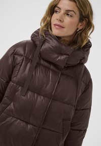 Tumši brūns polsterēts jakas, kurai ir augsta apkakle, priekšējās kabatas un eleganta apdare. Ietver regulējamas siksnas un kviltētu tekstūru.