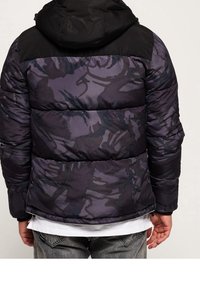 Vadderad jacka med svart huva och mörklila camouflage-mönster. Har kviltade sektioner och hög krage, tillverkad av ett slätt, syntetiskt material.