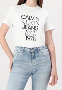 Camiseta de algodón blanca con texto gráfico negro "CALVIN KLEIN JEANS EST. 1978." Combinada con jeans de mezclilla de un azul claro con cintura alta y un ligero desgastado.