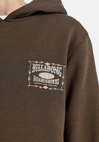 Brun hettegenser med et grafisk lapp med "Billabong Boardshorts"-logo. Teksten er i lyseblå og hvit, omgitt av en dekorativ kant.