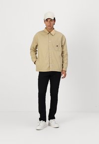 Beige skjortejakke med brystlomme, parret med sorte bukser og hvide sneakers. Modellen bærer en hvid cap.