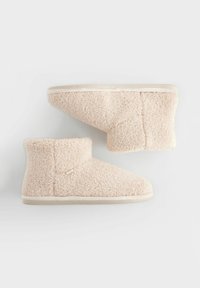 Chaussons beige en peluche avec un design mi-haut, une texture douce et un dessus simplement cousu avec une semelle de couleur claire.
