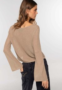 Suéter de punto beige con mangas acampanadas, con un escote ancho y un ajuste relajado, combinado con jeans negros adornados con tachuelas en los bolsillos.