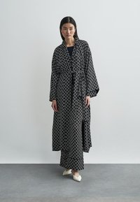 Μακριά μαύρη ρόμπα με λευκό γεωμετρικό μοτίβο, με φαρδιές μανσέτες και ζώνη που δένει στη μέση, φορεμένη πάνω από μαύρο τοπ και μακριά φούστα.