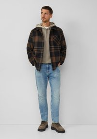 Karierte Flanelljacke in Dunkelbraun und Schwarz, über einem hellgrauen Hoodie getragen, kombiniert mit hellblauen Jeans und braunen Schnürstiefeln.