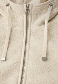 Sudadera con capucha de forro polar color crema, con una superficie texturizada, cierre de cremallera y cordón. Presenta herrajes plateados y una tela suave.