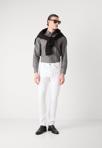 Siva košulja na dugmad, bijele slim-fit traperice, crni pulover prebačen preko ramena, crne sunčane naočale, sjajni crni mokasini, elegantan dizajn.