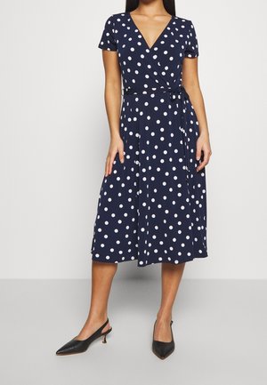 Nainen, joka pukeutunut tummansiniseen polvipituiseen kietaisumekkoon, jossa on valkoisia pilkkuja, ja mustoihin slingback-korkokenkiin, seisoo yksinkertaista vaaleaa taustaa vasten.