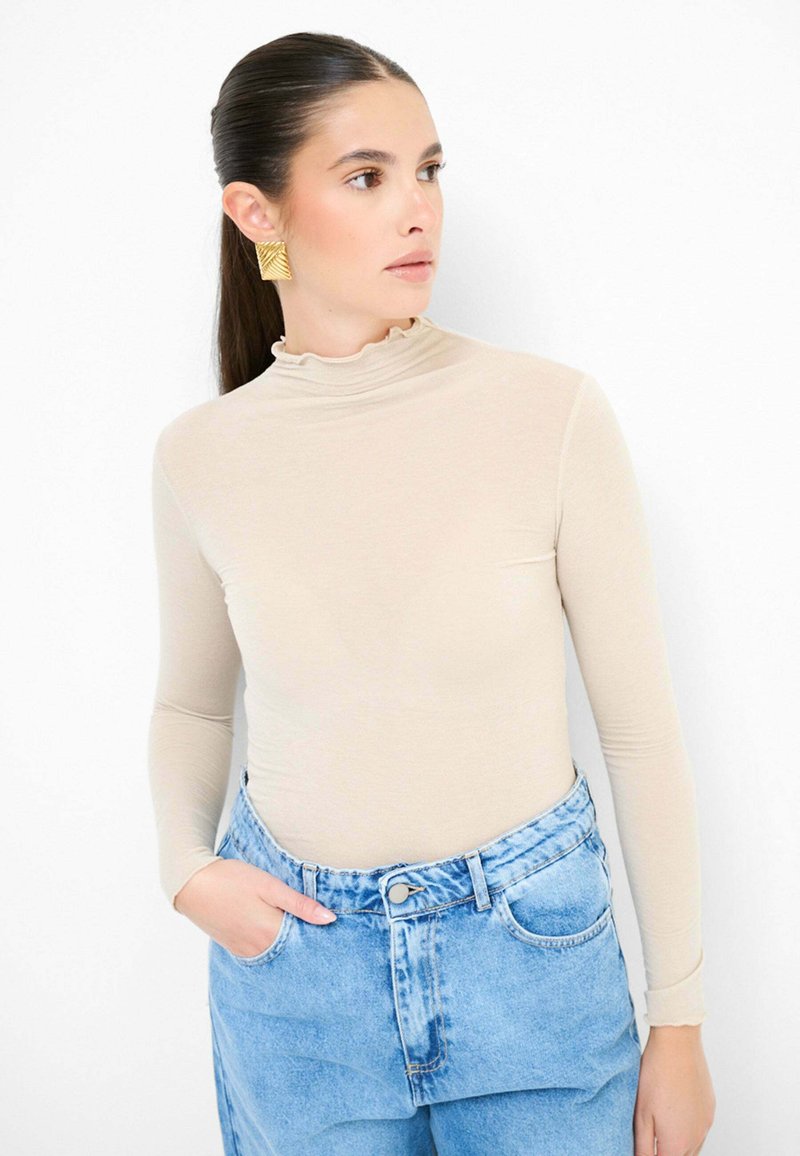 Top beige a maniche lunghe con scollo arricciato, abbinato a jeans in denim blu a vita alta. Il tessuto sembra liscio e aderente.