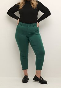 Vihreät istuvat housut, joissa on sileä pinta, lyhennetty pituus ja sivutaskut, yhdistetty mustaan ribbikangastoppiin pitkällä hihalla ja mustiin loafer-kenkiin.