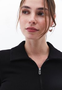 Musta ribaatu vetoketjullinen toppi laajalla kauluksella ja hopeisella vetoketjuyksityiskohdalla. Mallilla on luonnollinen meikki ja renkaankorvaket yhdistettynä valkoiseen taustaan.