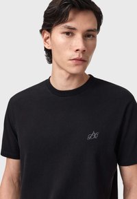 Μαύρο βαμβακερό T-shirt με στρογγυλή λαιμόκοψη, κοντά μανίκια και μικρό κεντητό λογότυπο στο στήθος. Λείο ύφασμα και μίνιμαλ  σχεδίαση.