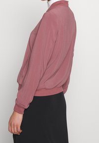 Veste bomber à manches longues avec col, en tissu doux rose poussiéreux. Comprend des poignets et un ourlet élastiques, des poches latérales et une texture lisse.