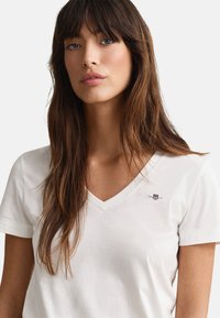 White cotton t-shirt with a V-neck and short sleeves, featuring a small embroidered logo on the left chest. Smooth texture.