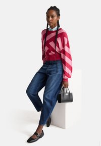 Rot- und rosa gestreifter Strickpullover, blaue Jeans, schwarze Ballerinas mit Schnalle, hält eine kleine strukturierte schwarze Handtasche, sitzt auf einem weißen Podest.