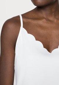 Primo piano di una persona che indossa un top senza maniche bianco con orlo a scallop, che mostra dettagli di spalle e clavicole.