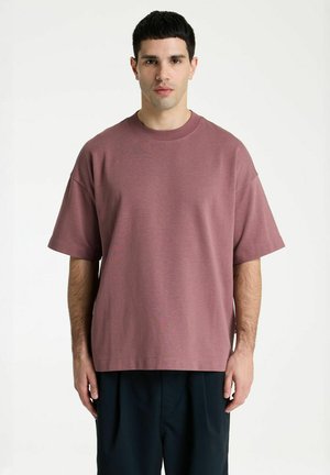 Junger Mann mit kurzen schwarzen Haaren, der ein lockeres mauvefarbenes T-Shirt und schwarze Hose trägt, steht vor einem schlichten weißen Hintergrund.