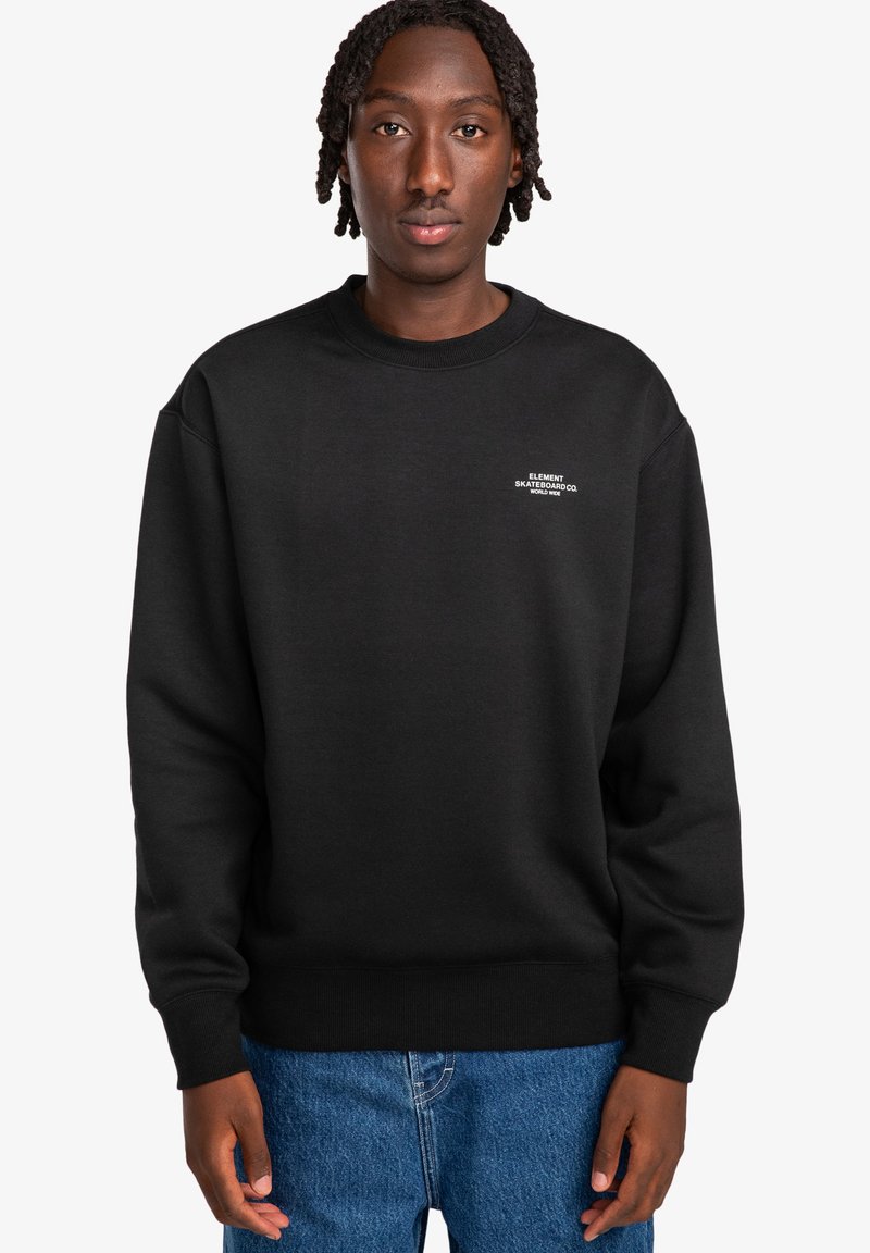 Felpa nera a girocollo in cotone, con un piccolo logo bianco sul petto, dotata di polsini e orlo a coste. Indossata con jeans in denim blu.