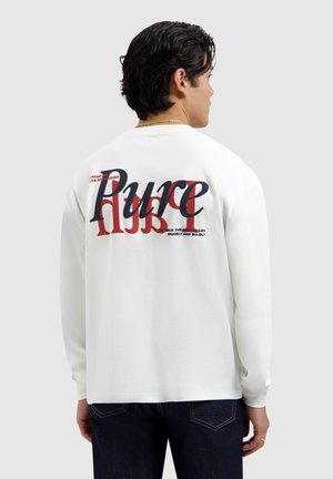Man draagt een wit shirt met lange mouwen met grote rode en zwarte tekst "Pure" en kleinere motiverende zinnen op de achterkant, donkere spijkerbroek, kijkt van zich af.