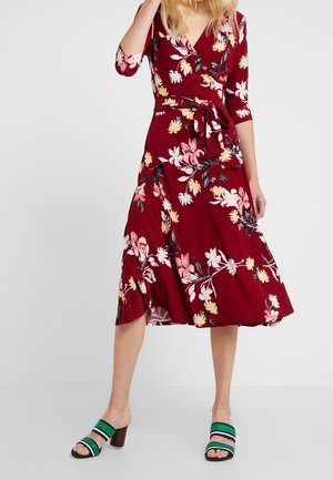 Frau trägt ein rotes, florales Wickelkleid mit dreiviertel Ärmeln und Gürtel in der Taille, kombiniert mit grün und marineblau gestreiften Sandalen mit Absatz.