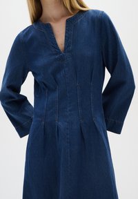 Denim haljina s dugim rukavima, V-izrezom i ušivenim detaljima. Ima usklađeni gornji dio i lepršavu suknju u srednje plavoj boji.