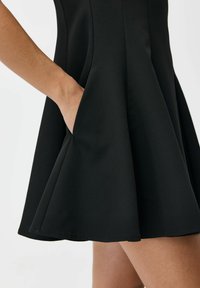 Dłoń umieszczona w bocznej kieszonce krótkiej, czarnej, rozszerzanej sukienki noszonej przez osobę o jasnym odcieniu skóry na białym tle.