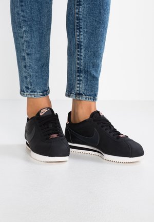 Pies con zapatillas Nike negras con detalles en rosa combinadas con vaqueros azules con puños, sobre un suelo y fondo blancos.